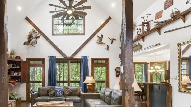 A spacious living room with a stone fireplace, a large ceiling fan, and a view of the outdoors through large windows. - Secluded 40 Acres Near Table Rock, Stunning Views, Games, Community Pool & Car Charger (Blue Eye)