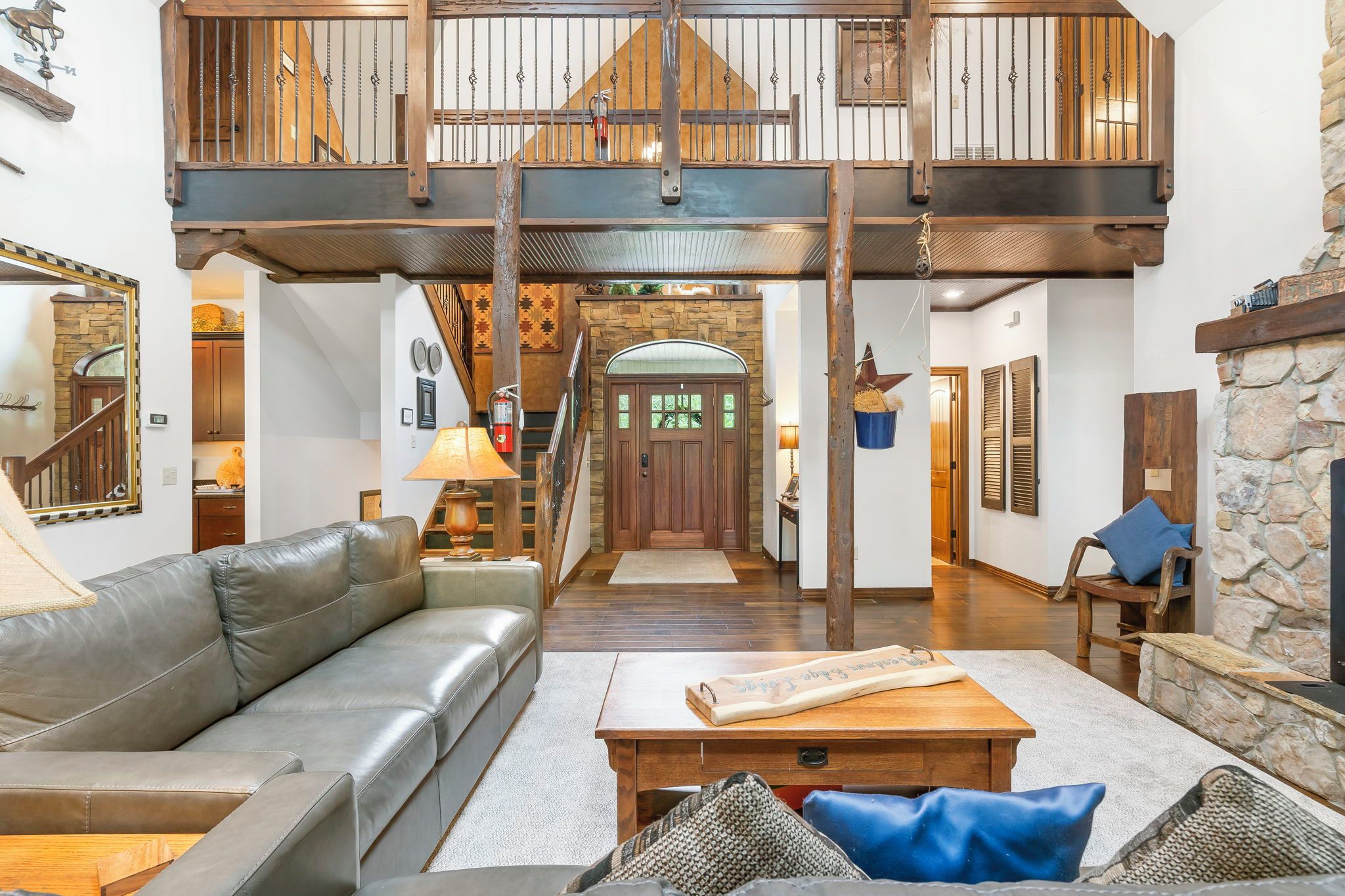 A spacious living area with a leather sofa, a wooden coffee table, and a stone fireplace. A staircase leads to an upper level with a balcony.