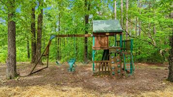 A wooden play structure with a swing set and climbing elements in a forested area.