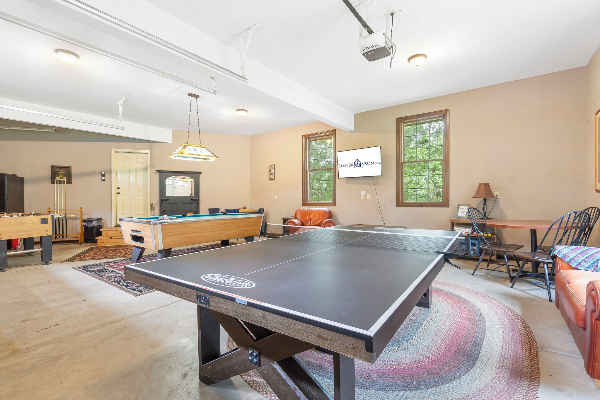 A spacious living room with a ping pong table, pool table, and a billiards table. There is a television mounted on the wall and a window providing natural light.