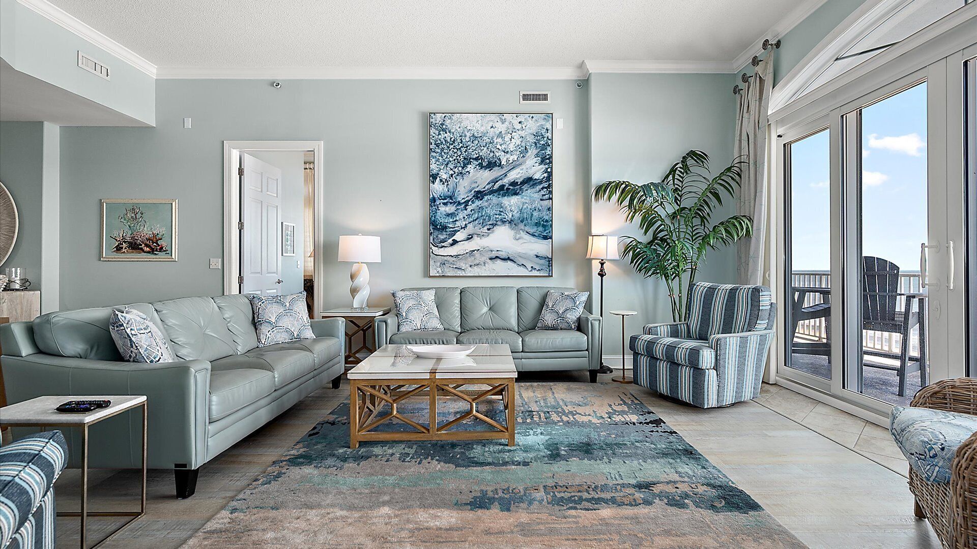 A modern living room with a grey couch, a coffee table, and a large abstract painting. There is a potted plant and a view of the outdoors through a sliding glass door.