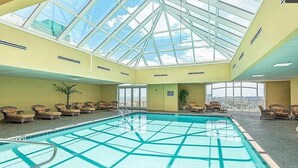An indoor swimming pool with a glass ceiling, lounge chairs, and a view of the city.
