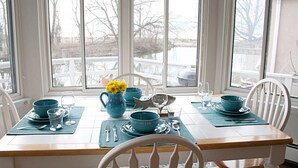 Una mesa de comedor dispuesta con vajilla azul y un florero con flores amarillas, ubicada cerca de amplios ventanales con vistas a un muelle y árboles.