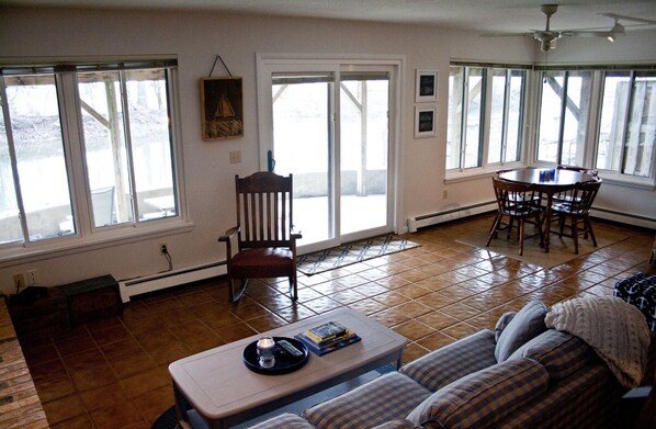 A living room with a sofa, a coffee table, a dining table with chairs, and a chair. There are large windows and a ceiling fan.