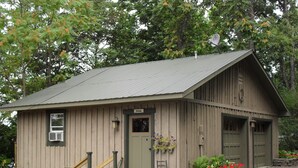 A rustic wooden building with a metal roof, a front door, and a small porch area with a chair and potted plants. - Cozy Cottage with 45' Lake Frontage and Kayaks (Lodi)