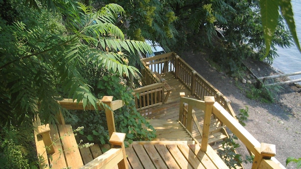 A wooden staircase leading to a wooden deck with railings, surrounded by lush greenery and a body of water in the background. - Cozy Cottage with 45' Lake Frontage and Kayaks (Lodi)