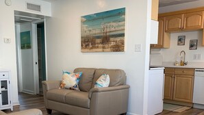 A modern living room with a beige sofa, wooden flooring, and a kitchen area with wooden cabinets and a white refrigerator. - Great Location ! 1 Short Block To The Boardwalk. (Virginia Beach)