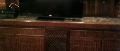 A bar area with a television, wooden cabinets, and shelves with various items.