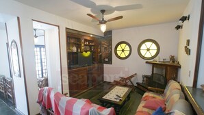 Ein Wohnzimmer mit einer Couch, einem Couchtisch und einem Deckenventilator. Ein Fenster mit Jalousien, eine Tür zu einem anderen Raum und eine Wand mit einer Uhr und einem Regal mit diversen Gegenständen.
