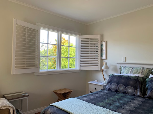 A bedroom with a bed, a nightstand, a bedside table, a window with blinds, and a small wooden stool.