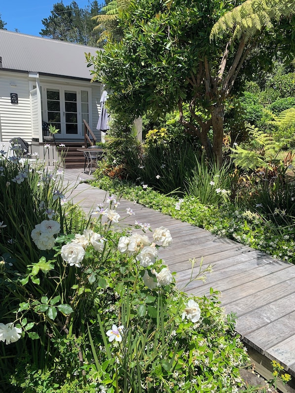 Una terraza de madera que conduce a una casa con porche, rodeada de abundante follaje y flores blancas.