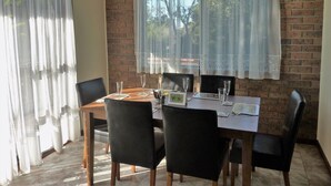 A dining area with a brick wall, wooden table, black chairs, and lace curtains. - Tura Views, 3 BR House (Tura Beach, Merimbula)
