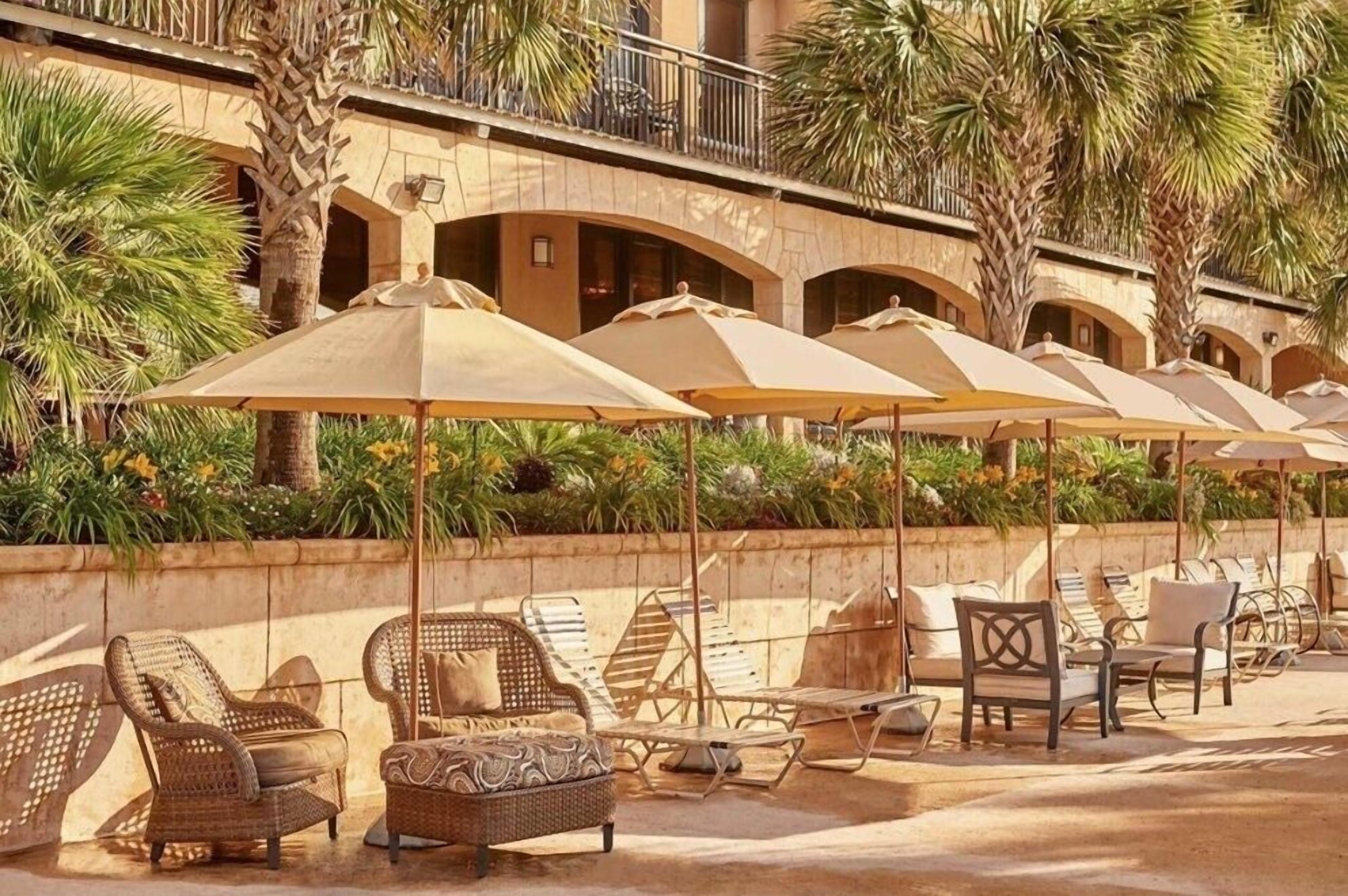 A sunlit outdoor seating area with lounge chairs and umbrellas, surrounded by palm trees and a building with arched balconies.