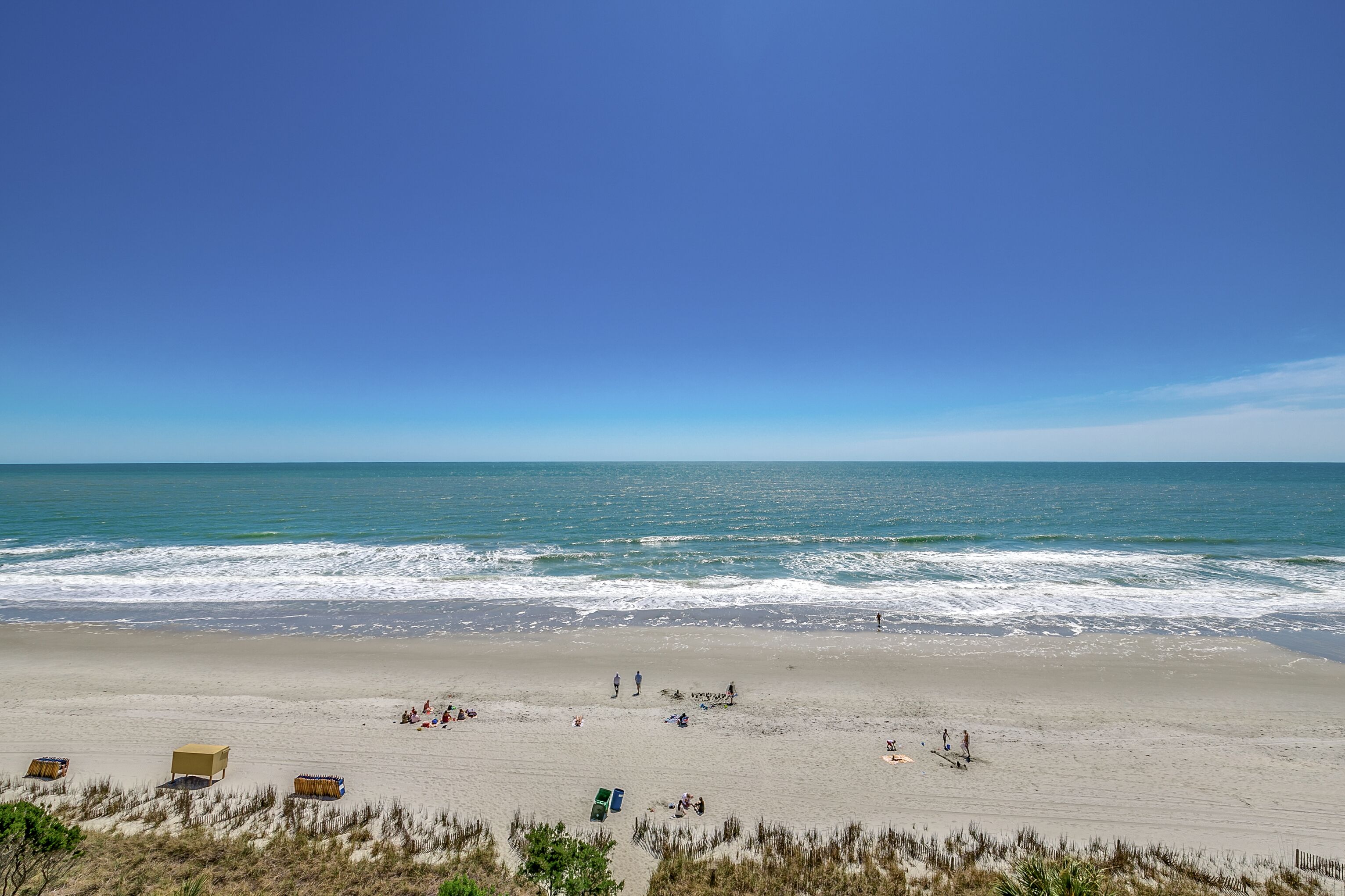 A beach with clear skies, calm waves, and a few people and beach huts.