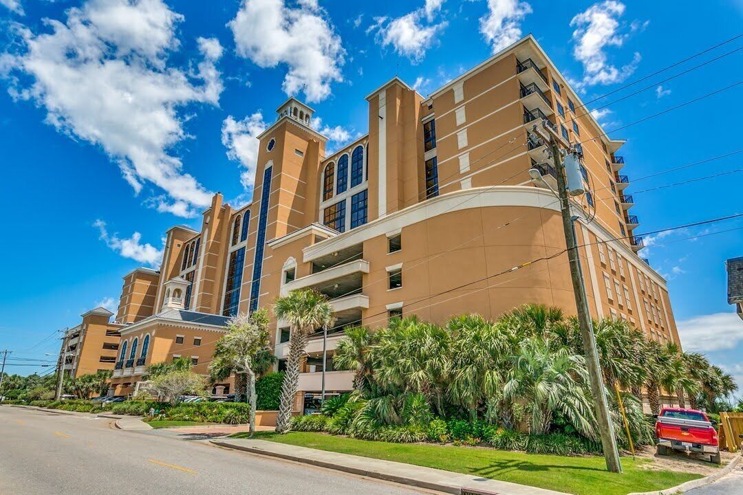 A multi-story hotel with a curved facade, balconies, and a parking area with a red vehicle.