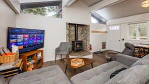 A modern living room with a flat-screen TV, grey sofas, a stone fireplace, and a dining area. - Lily Pad_Modern w/ AC! + Games, Smart TV's & HOA Amenities (Gym/HotTub/Sauna/Pool) (Truckee)