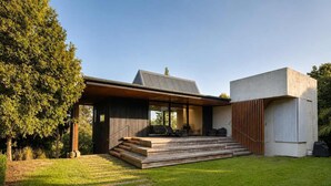 Uma casa moderna com um deck de madeira, área de estar coberta ao ar livre e uma grande árvore ao fundo.