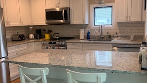 A modern kitchen with white cabinets, a granite countertop, a stainless steel refrigerator, a microwave, a coffee maker, and two white bar stools.