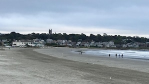 A wide sandy beach with a few people walking along the shore. In the background, there are houses and a church.