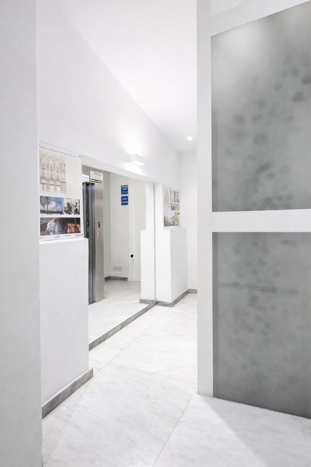 A modern, minimalist hallway with white walls and a grey accent wall, leading to a reception area with a digital display and a framed photo collage.
