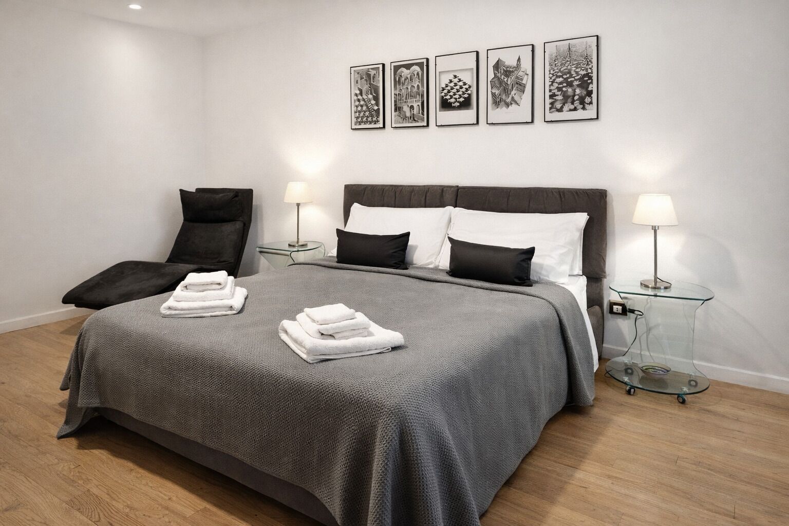 Une chambre à coucher moderne avec un grand lit, deux tables de chevet avec des lampes, un fauteuil et un mur décoré de tableaux encadrés.