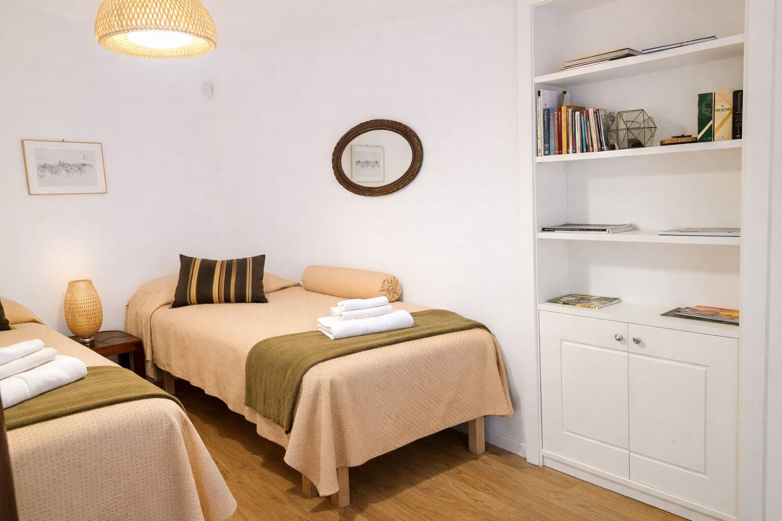A hotel room with two beds, a bookshelf, and a wall-mounted mirror.