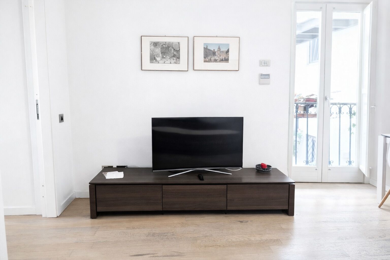 A modern living room with a flat-screen TV on a dark wooden cabinet, white walls, and two framed pictures.