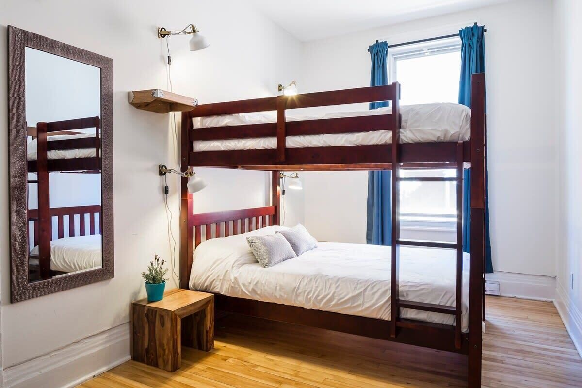 A bunk bed room with a wooden floor, a small wooden table with a plant, a mirror, and a window with curtains.