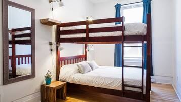 A bunk bed room with a wooden floor, a small wooden table with a plant, a mirror, and a window with curtains.