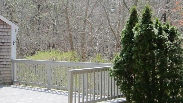 A wooden deck with a railing, surrounded by dense trees and shrubbery. - Quaint 2BR Cottage in Wellfleet (1724) (Wellfleet)
