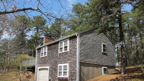 A two-story house with a garage, surrounded by trees. - Quaint 2BR Cottage in Wellfleet (1724) (Wellfleet)