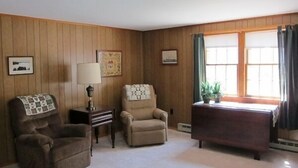 A cozy room with wooden paneling, a brown armchair, a beige armchair, a small table with a lamp, and a desk with a plant. - Quaint 2BR Cottage in Wellfleet (1724) (Wellfleet)