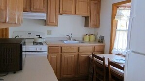 A compact kitchen with wooden cabinets, a stove, a sink, and a dining area with a table and chairs. - Quaint 2BR Cottage in Wellfleet (1724) (Wellfleet)
