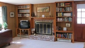 A cozy living room with a fireplace, wooden bookshelves, and a television. - Quaint 2BR Cottage in Wellfleet (1724) (Wellfleet)