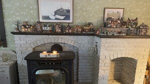 A cozy room with a stone fireplace, a small table with candles, and a mantelpiece decorated with miniature houses and a Christmas tree. - Mount Desert Island, Bar Harbor, Acadia National Park (Bar Harbor)