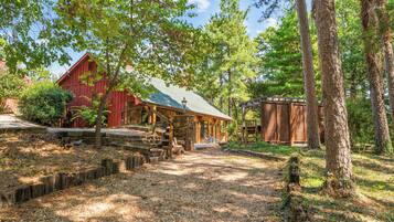 Une cabane rustique avec des fondations en pierre, une véranda en bois et, au loin, une grange rouge.