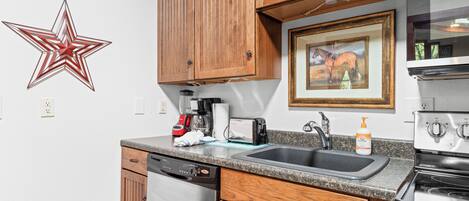 A kitchen with wooden cabinets, a dishwasher, a coffee maker, a toaster, a sink, and a framed picture on the wall.
