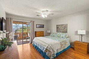 Une chambre à coucher avec un lit, un ventilateur de plafond, une table de chevet, une lampe, un téléviseur et une vue sur l’extérieur.