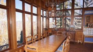 A large wooden dining table with benches, a chandelier, and a view of trees through large windows.