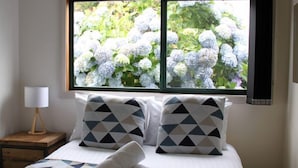 Une chambre à coucher avec un lit, des oreillers, une lampe et une fenêtre donnant sur des hortensias.
