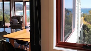 Une pièce dotée d’une grande fenêtre donnant sur la mer et les collines. On y trouve une table et des chaises en bois, ainsi qu’un fauteuil à motifs.
