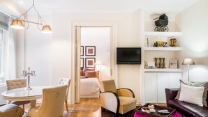 Een moderne hotelkamer met een ronde eettafel, twee fauteuils, een bank, een aan de muur bevestigde televisie en een klein boekenrek met decoratieve objecten.