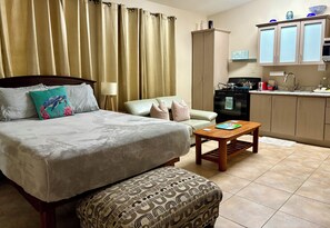 Quarto de hotel com cama, sofá, um banquinho estampado, uma mesa pequena, cozinha com armários e eletrodomésticos, e janela com cortinas.