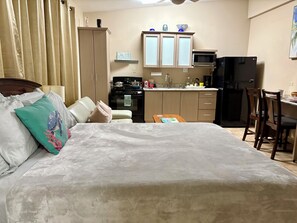 Quarto com cama, mesa de cabeceira e vista para uma cozinha com armários, micro-ondas e geladeira.