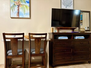 Quarto com televisão sobre um suporte de madeira, duas cadeiras de madeira, cômoda de madeira com gavetas e um quadro de uma palmeira na parede.