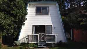 A white, two-story house with a steeply pitched roof, a front porch with a wooden bench, and a small window. - Kiwi Bach by the Beach (Castle Point)
