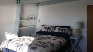 A bedroom with a bed, a bedside lamp, and a shelf with a picture frame. - Kiwi Bach by the Beach (Castle Point)