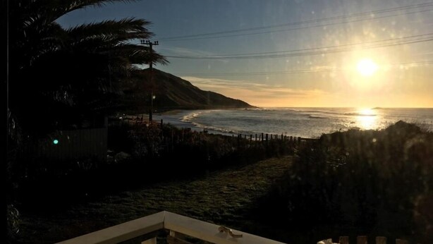 A sunset view from a balcony overlooking a beach with palm trees and a fence. - Kiwi Bach by the Beach (Castle Point)