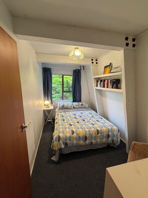 A bedroom with a bed, a desk, a chair, a bookshelf, and a window with curtains. - Kiwi Bach by the Beach (Castle Point)