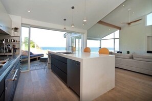 A modern kitchen with a large island, a dining area with a view, and a living space with a sofa and a plant. - Northern Beaches beachfront house . (Warriewood)